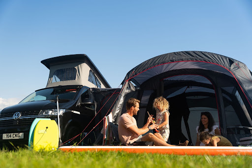 An image of a black campervan with an awning attached. There is a family of four sitting inside.