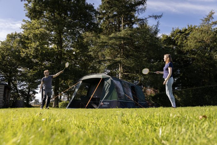 Two individuals playing tennis on a grassy field, with a tent set up in the background.
