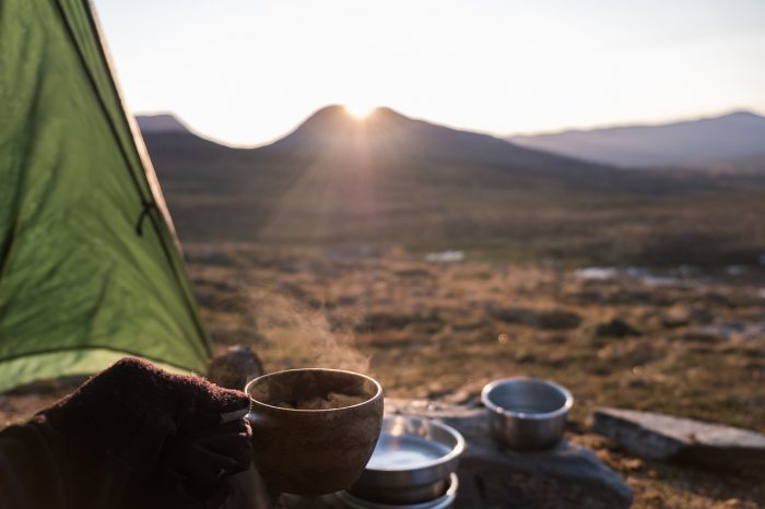 A person enjoys a cup of coffee while sitting beside a tent in a serene outdoor setting.