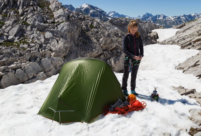 A woman stands beside a tent on a snowy mountain, surrounded by a serene winter landscape.