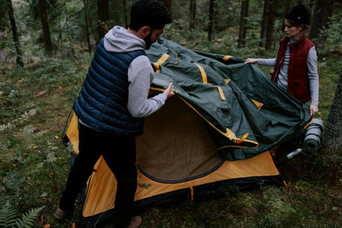 Two individuals assembling a tent amidst a lush forest setting, surrounded by trees and natural greenery.