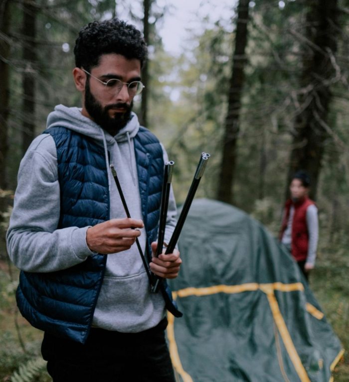 A man stands in the woods assembling tent poles, surrounded by trees and foliage.
