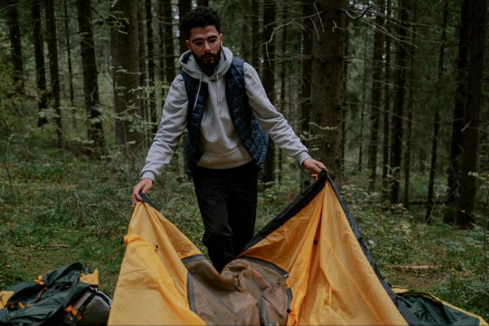 A man lays out a tent in a wooded area, surrounded by trees and natural scenery.