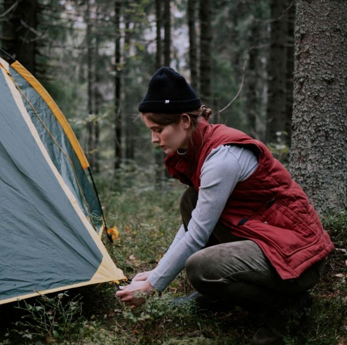 A woman wearing a red vest and black hat is setting up a tent outdoors.