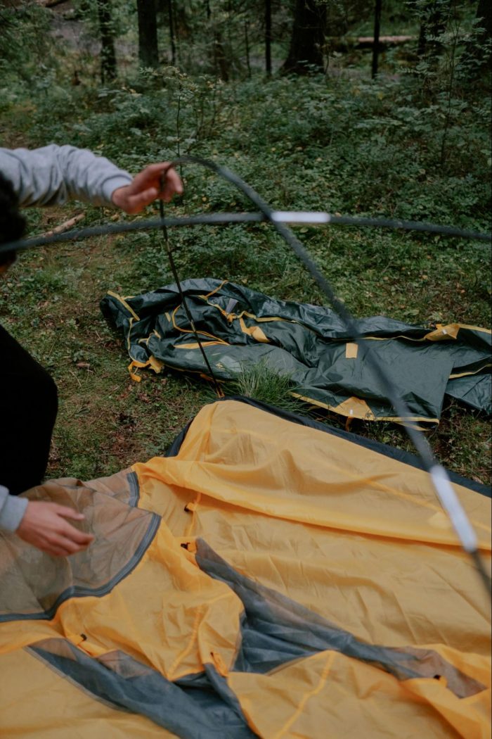 A man is assembling tent poles in a wooded area, surrounded by trees and natural scenery.