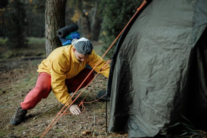 A man wearing a yellow jacket and red pants is tensioning tent guylines in a natural setting.