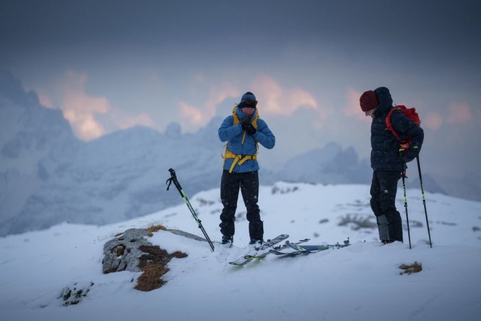 Two individuals standing triumphantly on a snow-covered mountain peak, surrounded by a clear blue sky.
