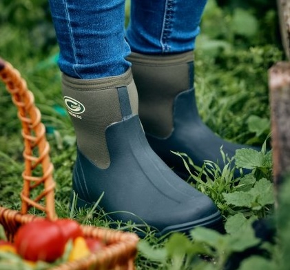 A person in rubber boots stands in grass, holding a basket filled with fresh vegetables.
